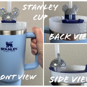 May include: A light blue Stanley travel mug with a black bear logo and the text "Stanley Since 1913". The mug has a handle and a white lid with a silver glitter Mickey Mouse ear lid topper and a straw.