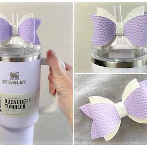 May include: A white stainless steel tumbler with a purple and white bow on the lid. The tumbler has a black and white label that says "Stanley The Flowstate Quencher H2.0 Tumbler 40 oz | 1.18 L Limited Edition Color"