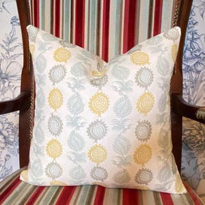 May include: A square cushion featuring a repeating pattern of yellow, grey, and pale blue floral motifs on a white background. The cushion is placed on a striped chair with red, white, and blue stripes.