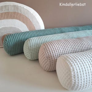 Op de afbeelding: Een verzameling cilindrische kussens in verschillende kleuren, waaronder teal, lichtgroen, beige en wit, met een wafelstructuur. Een regenboogvormig kussen in bijpassende kleuren is ook zichtbaar. De kussens zijn op een wit oppervlak geplaatst.