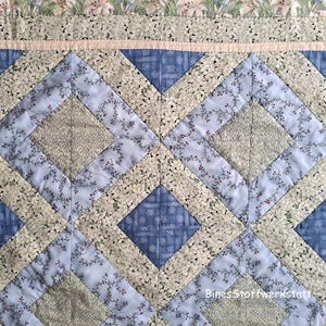 May include: Close-up of a quilted fabric with a diamond pattern. The quilt features a combination of blue, green, and cream-colored floral and geometric patterns. The design includes a border with floral patterns and a light pink strip.