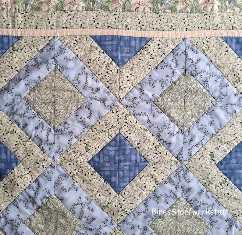 May include: Close-up of a quilted fabric with a diamond pattern. The quilt features a combination of blue, green, and cream-colored floral and geometric patterns. The design includes a border with floral patterns and a light pink strip.