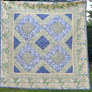 May include: A handmade quilt with a central geometric pattern in shades of blue and green. The quilt features a floral border with white lilies on a light green background. The text "BinesStoffwerkstatt" is visible in the lower right corner.
