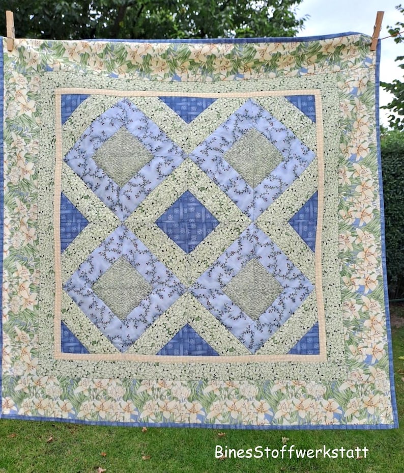 May include: A handmade quilt with a central geometric pattern in shades of blue and green. The quilt features a floral border with white lilies on a light green background. The text "BinesStoffwerkstatt" is visible in the lower right corner.