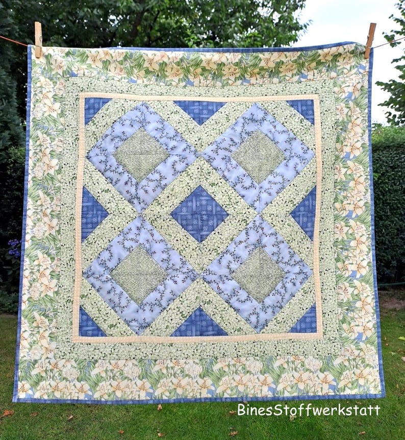 May include: A square quilt with a floral border and a geometric center design. The center features blue and green diamond shapes. The border has a cream background with white and yellow flowers. The quilt is hanging on a clothesline.