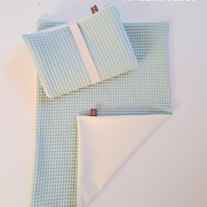 Könnte beinhalten: Ein Set aus hellblauem Waffelstoff, bestehend aus einer rechteckigen Decke und einem kleineren quadratischen Artikel, möglicherweise ein Kissen oder eine Unterlage. Ein weißes Band sichert den kleineren Artikel. Der Text "Kindallerliebst" ist oben rechts sichtbar.