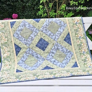 May include: A handmade quilt with a geometric design in shades of blue and green. The quilt features a central pattern of interlocking squares and diamonds, with a floral border. The text "BinesStoffwerkstatt" is visible in the upper right corner.