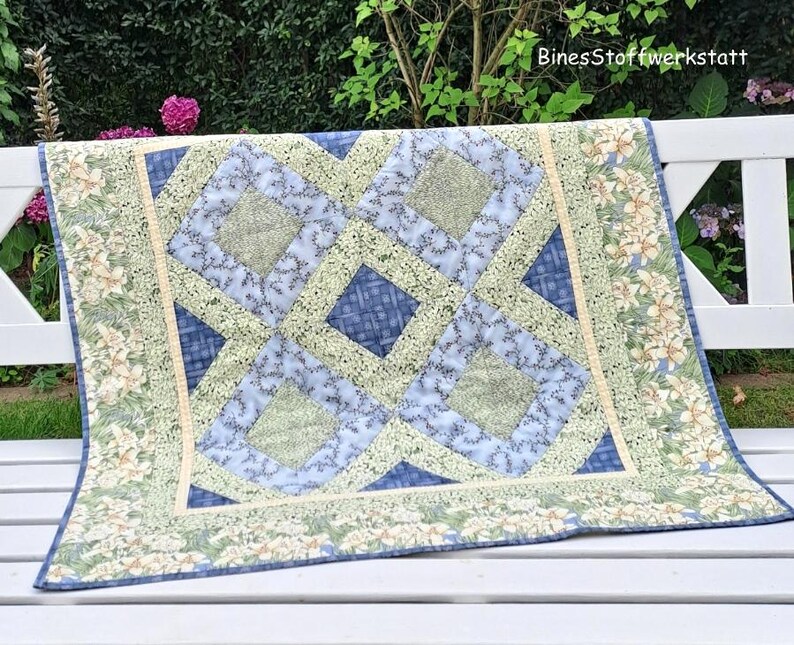 May include: A handmade quilt with a geometric design in shades of blue and green. The quilt features a central pattern of interlocking squares and diamonds, with a floral border. The text "BinesStoffwerkstatt" is visible in the upper right corner.