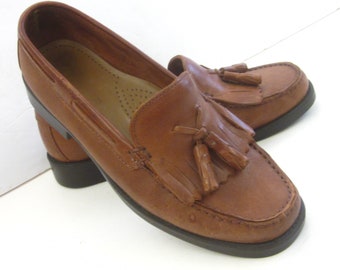 May include: Brown leather tassel loafers with a black rubber sole. The loafers have a slip-on design and a tassel detail on the front.