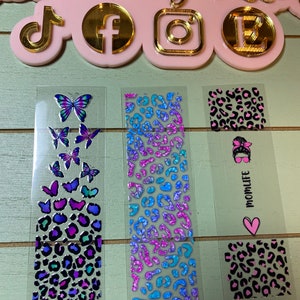 May include: Three clear sticker sheets with various designs. The first sheet features a stack of colorful butterflies. The second sheet has a repeating pattern of blue, pink, and purple leopard print. The third sheet has a pink heart with the text "MOMLIFE" and a leopard print design.