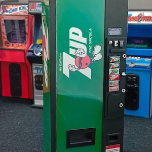 Può includere: Un distributore automatico di bevande in miniatura verde e nero con il logo 7UP, la scritta The Uncola e una selezione di pulsanti per bevande tra cui 7UP, Dr Pepper, Mountain Dew e Sunkist. La parte anteriore include una fessura e un'etichetta.