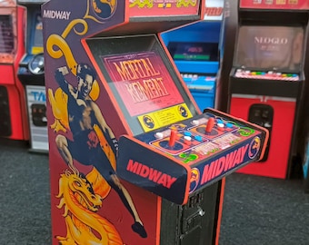 Op de afbeelding: Een miniatuur Mortal Kombat-arcadekast met een rood en zwart zijpaneel met een afbeelding van een vechter en een gele draak. Het scherm toont het Mortal Kombat-logo en het bedieningspaneel bevat twee rode joysticks en knoppen.