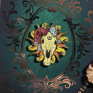 May include: A gold enamel pin featuring a skull with a third eye, surrounded by flowers and a sunburst design. The pin is on a dark green background with gold foil accents and the text "Wise Words" and "Kantaos".