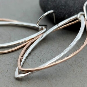 May include: A pair of silver and rose gold earrings with a delicate, curved design. The earrings are made of two thin wires that are intertwined and have a hammered texture.