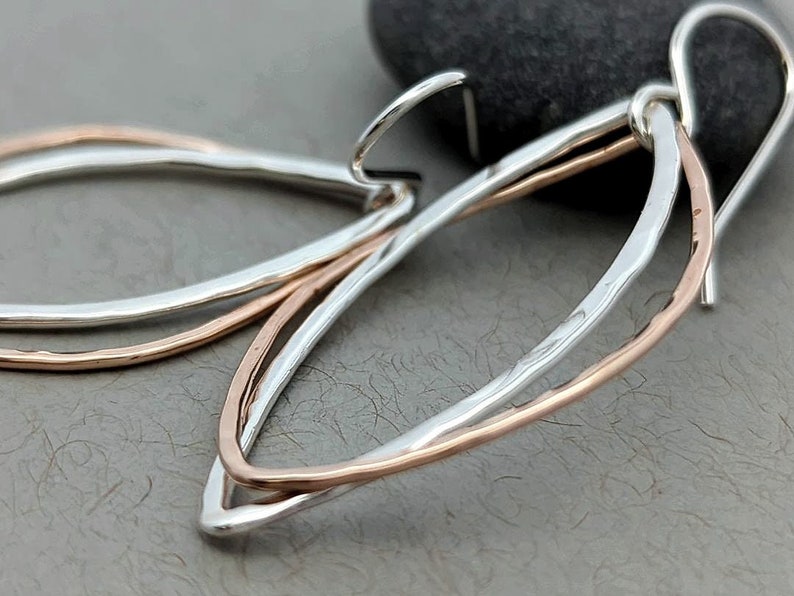 May include: A pair of silver and rose gold earrings with a delicate, curved design. The earrings are made of two thin wires that are intertwined and have a hammered texture.