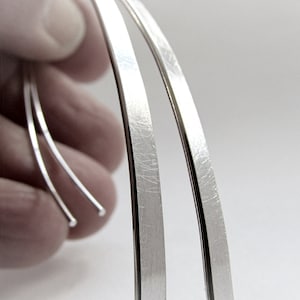 May include: A pair of silver-colored earrings with a minimalist design. The earrings have a curved, elongated shape, with a brushed metal finish. The image is set against a white background, highlighting the jewelry's sleek lines.
