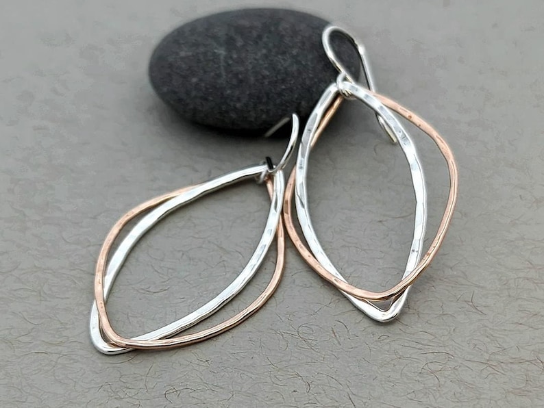 May include: A pair of silver and rose gold earrings with a hammered finish. The earrings are shaped like an oval with a smaller oval inside.