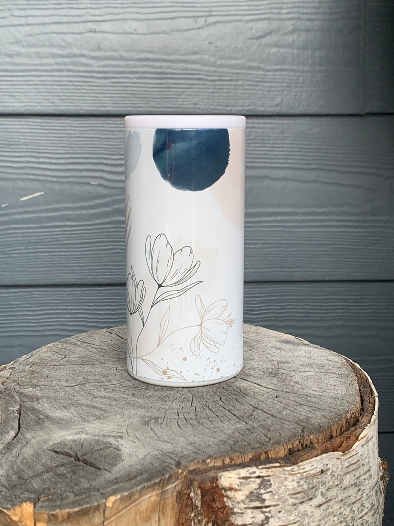 Puede incluir: Un vaso de acero inoxidable blanco con un dise&ntilde;o floral en negro y dorado. El dise&ntilde;o presenta formas abstractas en azul y rosa.
