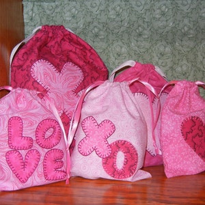 May include: Four pink fabric drawstring bags with different designs. The bags have pink ribbon ties and feature hearts, the words "LOVE" and "XO" in pink fabric.
