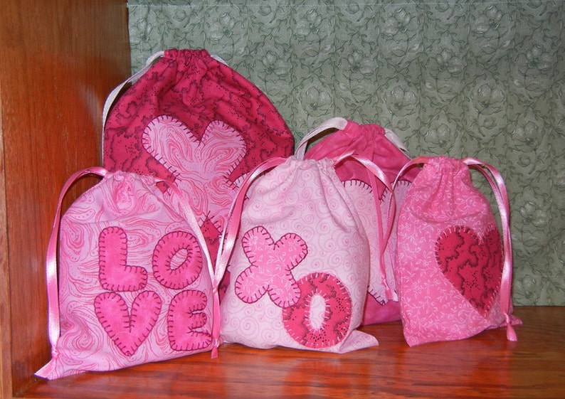 May include: Four pink fabric drawstring bags with different designs. The bags have pink ribbon ties and feature hearts, the words "LOVE" and "XO" in pink fabric.