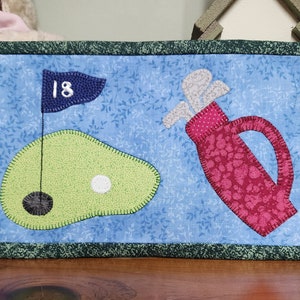 Peut inclure: Une suspension murale matelass&eacute;e bleue et verte avec un sac de golf, une balle de golf et un drapeau de golf avec le num&eacute;ro 18 dessus.