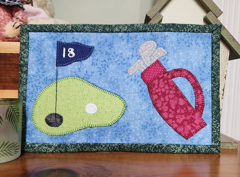 Peut inclure: Une suspension murale matelass&eacute;e bleue et verte avec un sac de golf, une balle de golf et un drapeau de golf avec le num&eacute;ro 18 dessus.
