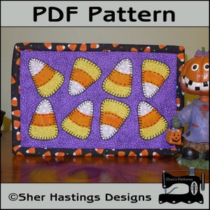 Puede incluir: Un edredón rectangular con temática de Halloween con ocho diseños de caramelos de maíz sobre un fondo morado. El edredón está bordeado con una tela negra adornada con caramelos de maíz. El texto "PDF Pattern" está en la parte superior.
