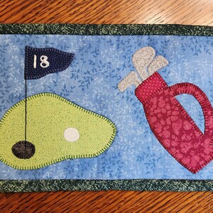 Peut inclure: Un dessous de verre matelass&eacute; bleu et vert avec un sac de golf et un green de golf avec le num&eacute;ro 18 sur un drapeau.