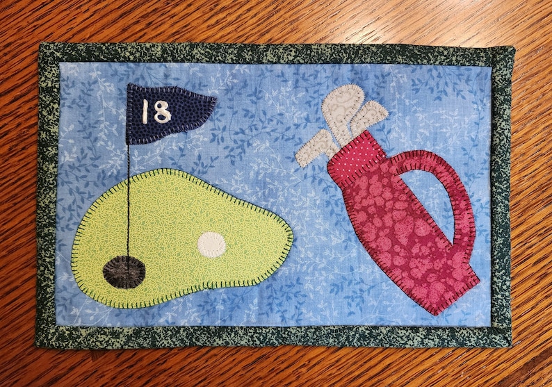 Peut inclure: Un dessous de verre matelass&eacute; bleu et vert avec un sac de golf et un green de golf avec le num&eacute;ro 18 sur un drapeau.