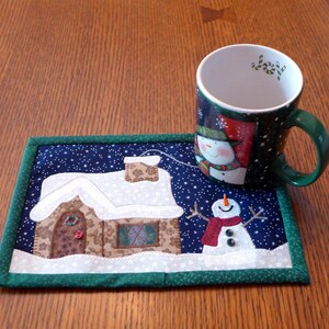 K&ouml;nnte beinhalten: Eine gr&uuml;ne und wei&szlig;e Tasse mit Weihnachtsmotiv und einem Schneemann-Design steht auf einem gesteppten Untersetzer mit einem verschneiten Haus und einem Schneemann-Design. Die Tasse hat das Wort "Joy" auf der Innenseite.
