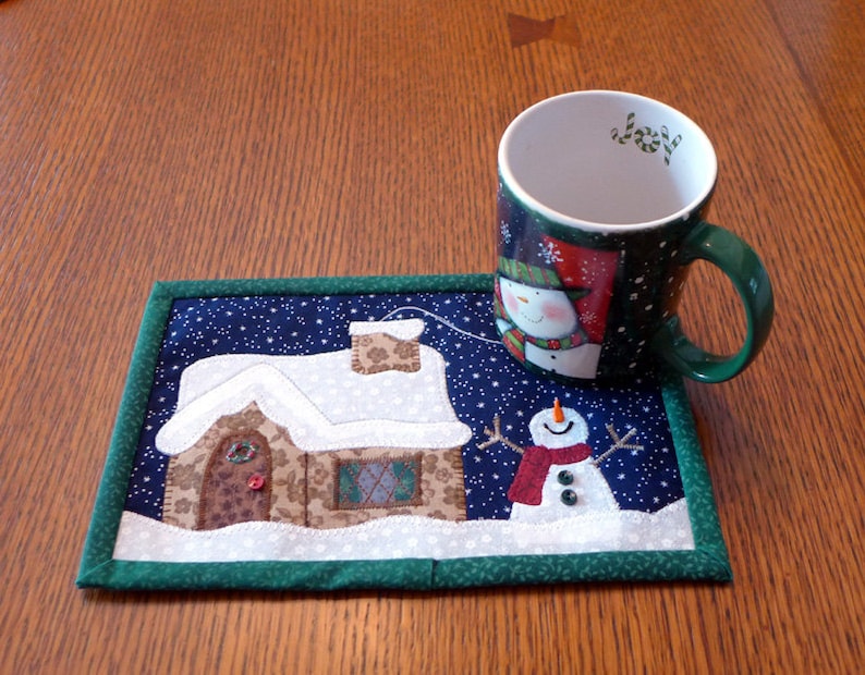 K&ouml;nnte beinhalten: Eine gr&uuml;ne und wei&szlig;e Tasse mit Weihnachtsmotiv und einem Schneemann-Design steht auf einem gesteppten Untersetzer mit einem verschneiten Haus und einem Schneemann-Design. Die Tasse hat das Wort "Joy" auf der Innenseite.
