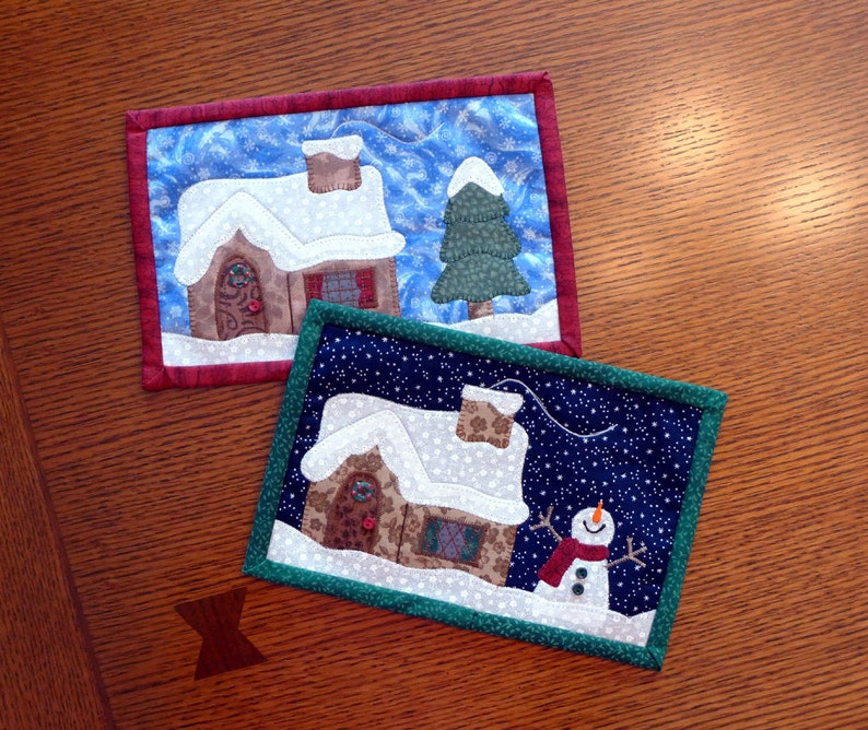 K&ouml;nnte beinhalten: Zwei gesteppte Untersetzer zum Thema Weihnachten. Der erste Untersetzer zeigt ein braunes Haus mit einem wei&szlig;en Dach und einem gr&uuml;nen Baum. Der zweite Untersetzer zeigt ein braunes Haus mit einem wei&szlig;en Dach und einen Schneemann.