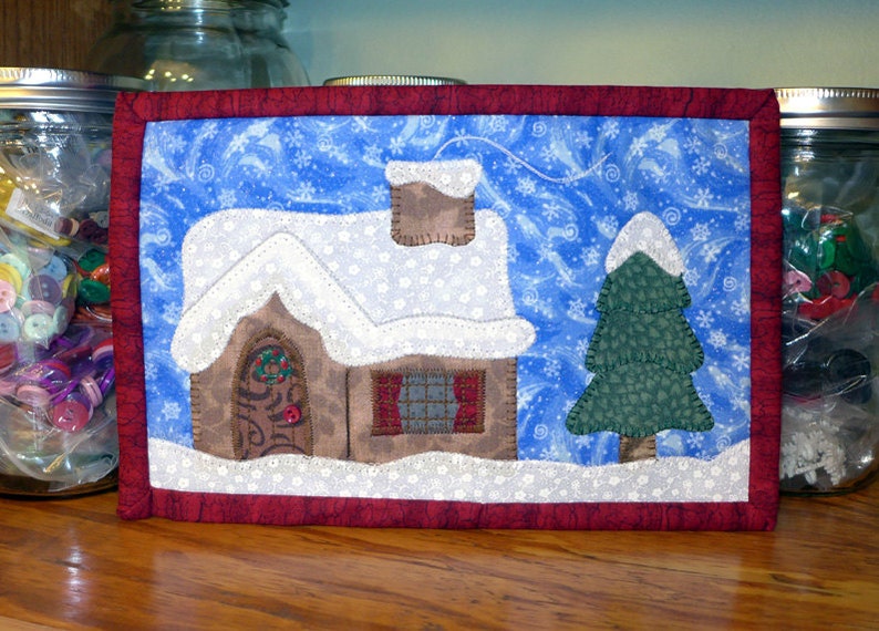 K&ouml;nnte beinhalten: Ein gesteppter Wandbehang mit einem schneebedeckten Haus und einem Kiefernbaum auf blauem Hintergrund mit wei&szlig;en Schneeflocken. Das Haus hat eine braune T&uuml;r mit einem Kranz und ein Fenster. Der Baum ist gr&uuml;n mit einer wei&szlig;en Schneehaube.