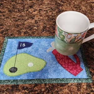 Peut inclure: Un dessous de tasse en tissu bleu et vert sur le th&egrave;me du golf avec un golfeur et un green avec un drapeau qui indique "18". Une tasse blanche avec un motif floral vert, jaune et orange est pos&eacute;e sur le dessous de tasse.