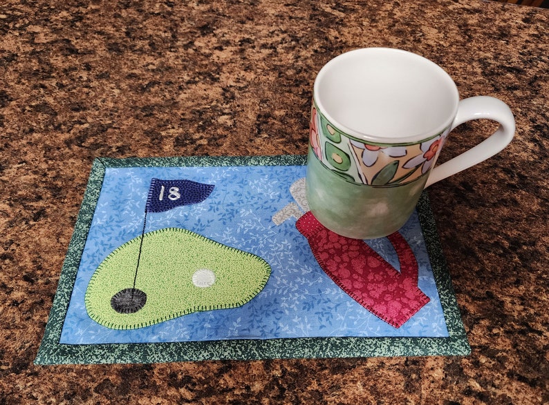 Peut inclure: Un dessous de tasse en tissu bleu et vert sur le th&egrave;me du golf avec un golfeur et un green avec un drapeau qui indique "18". Une tasse blanche avec un motif floral vert, jaune et orange est pos&eacute;e sur le dessous de tasse.