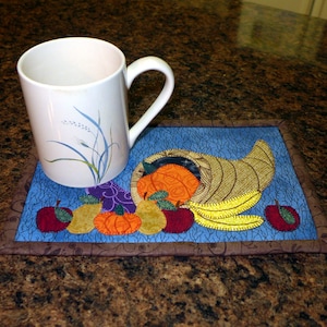 May include: A blue and brown quilted coaster with a cornucopia design featuring a variety of fruits and vegetables, including apples, pears, pumpkins, and a banana. A white mug with a floral design sits on top of the coaster.