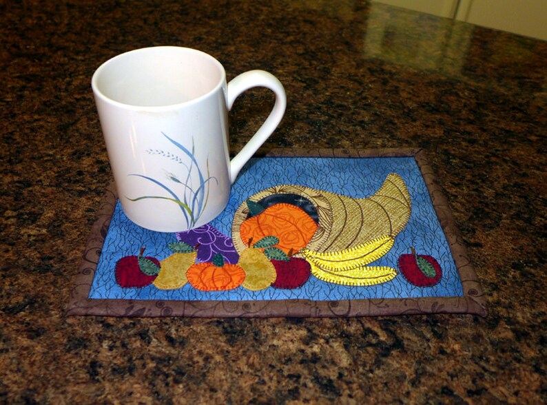 May include: A blue and brown quilted coaster with a cornucopia design featuring a variety of fruits and vegetables, including apples, pears, pumpkins, and a banana. A white mug with a floral design sits on top of the coaster.