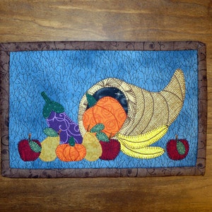 May include: A quilted wall hanging with a cornucopia overflowing with fall harvest fruits and vegetables. The cornucopia is brown and gold, and the fruits and vegetables include apples, pears, pumpkins, and an eggplant. The background is a blue fabric with a textured pattern.