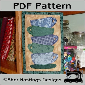 May include: A PDF pattern for a quilted wall hanging featuring a stack of five teacups in shades of blue and green. The teacups are decorated with different patterns, including floral, plaid, and polka dots. The pattern is framed with a green border and includes the text "Sher Hastings Designs".