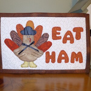 Peut inclure: Une suspension murale matelass&eacute;e marron et blanche avec une applique de dinde et le texte "EAT HAM".