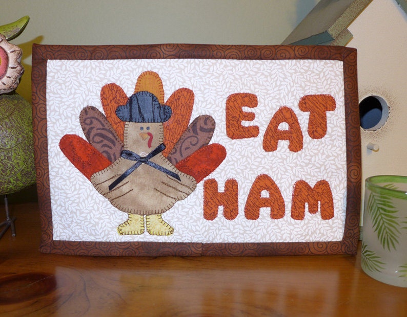 Peut inclure: Une suspension murale matelass&eacute;e marron et blanche avec une applique de dinde et le texte "EAT HAM".