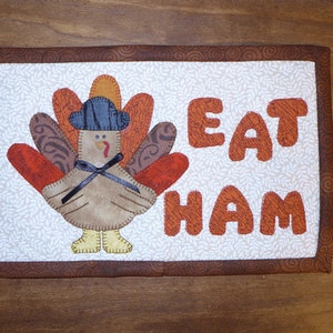 Peut inclure: Un panneau en tissu matelass&eacute; brun et blanc avec une dinde portant un chapeau bleu et les mots "Eat Ham" en lettres orange.