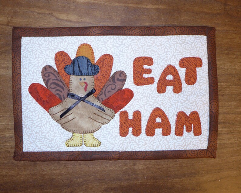 Peut inclure: Un panneau en tissu matelass&eacute; brun et blanc avec une dinde portant un chapeau bleu et les mots "Eat Ham" en lettres orange.