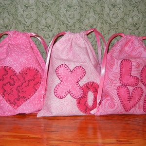 May include: Three pink fabric drawstring bags with different Valentine's Day themed appliques. The bags are decorated with a heart, an 'XO' and the word 'LOVE'.