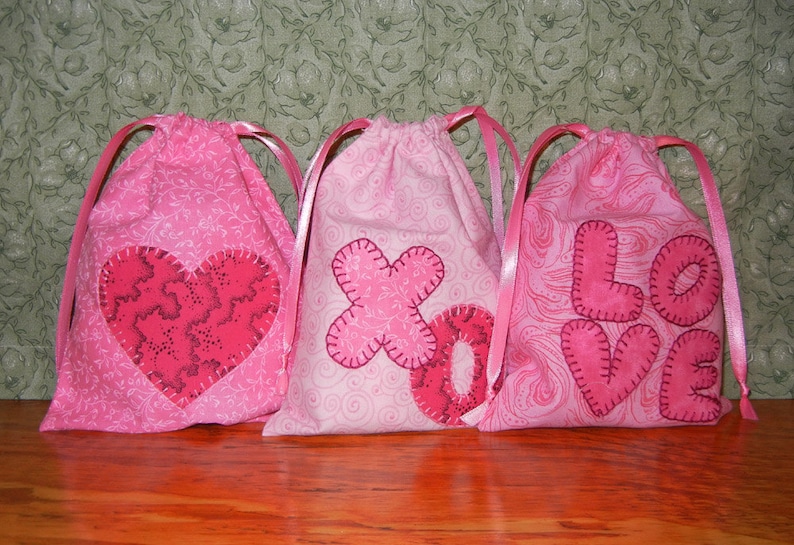 May include: Three pink fabric drawstring bags with different Valentine's Day themed appliques. The bags are decorated with a heart, an 'XO' and the word 'LOVE'.