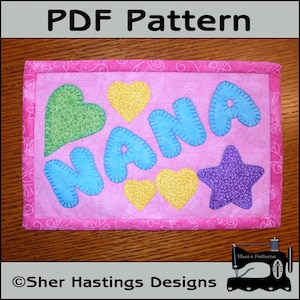 May include: A pink fabric square with a stitched border and the word "NANA" in blue, yellow, and green letters. There are also hearts and a star in the design. The fabric square is on a wooden surface.
