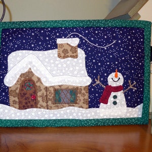 K&ouml;nnte beinhalten: Ein gesteppter Wandbehang mit einer Winterszene. Ein braunes Lebkuchenhaus mit einem wei&szlig;en Dach und ein Schneemann mit rotem Schal stehen vor einem dunkelblauen Hintergrund mit wei&szlig;en Schneeflocken.