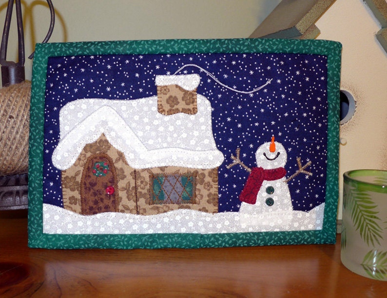 K&ouml;nnte beinhalten: Ein gesteppter Wandbehang mit einer Winterszene. Ein braunes Lebkuchenhaus mit einem wei&szlig;en Dach und ein Schneemann mit rotem Schal stehen vor einem dunkelblauen Hintergrund mit wei&szlig;en Schneeflocken.