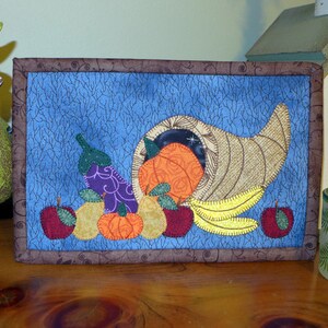 May include: A framed quilted wall hanging with a cornucopia overflowing with fruit and vegetables. The cornucopia is brown and gold, the fruit is red, orange, and yellow, and the vegetables are purple and green. The background is a blue fabric with a white, brown, and black pattern.
