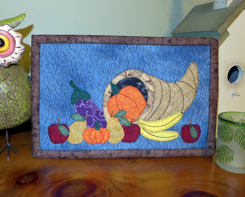May include: A framed quilted wall hanging with a cornucopia overflowing with fruit and vegetables. The cornucopia is brown and gold, the fruit is red, orange, and yellow, and the vegetables are purple and green. The background is a blue fabric with a white, brown, and black pattern.